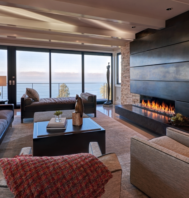 A modern living room with a large fireplace, a sofa, and a daybed positioned in front of floor-to-ceiling windows and more overlooking a lake.