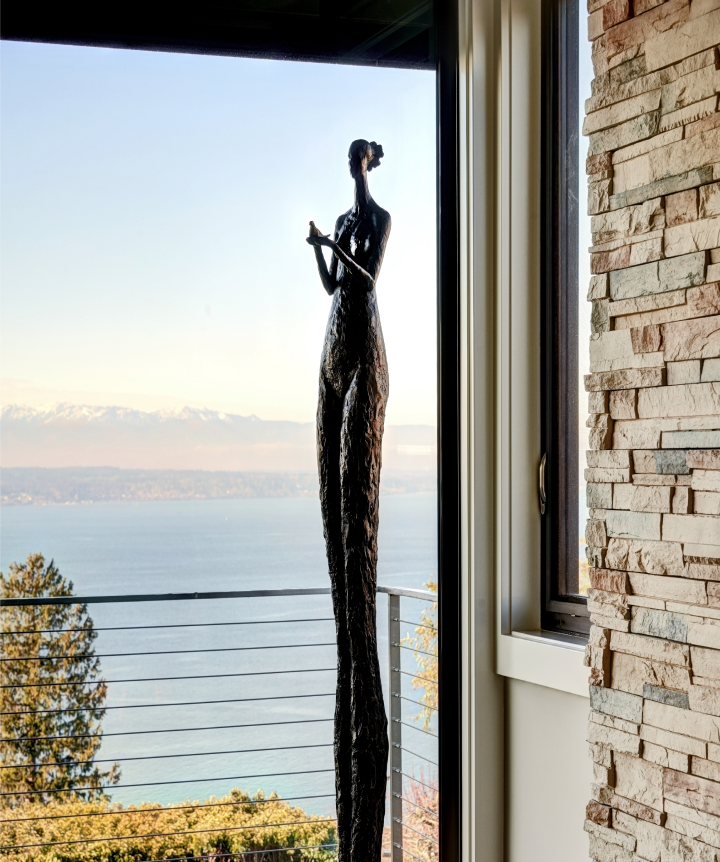 A tall, slender bronze sculpture of a woman stands before windows, doors and more that overlook a body of water and distant snow-capped mountains.