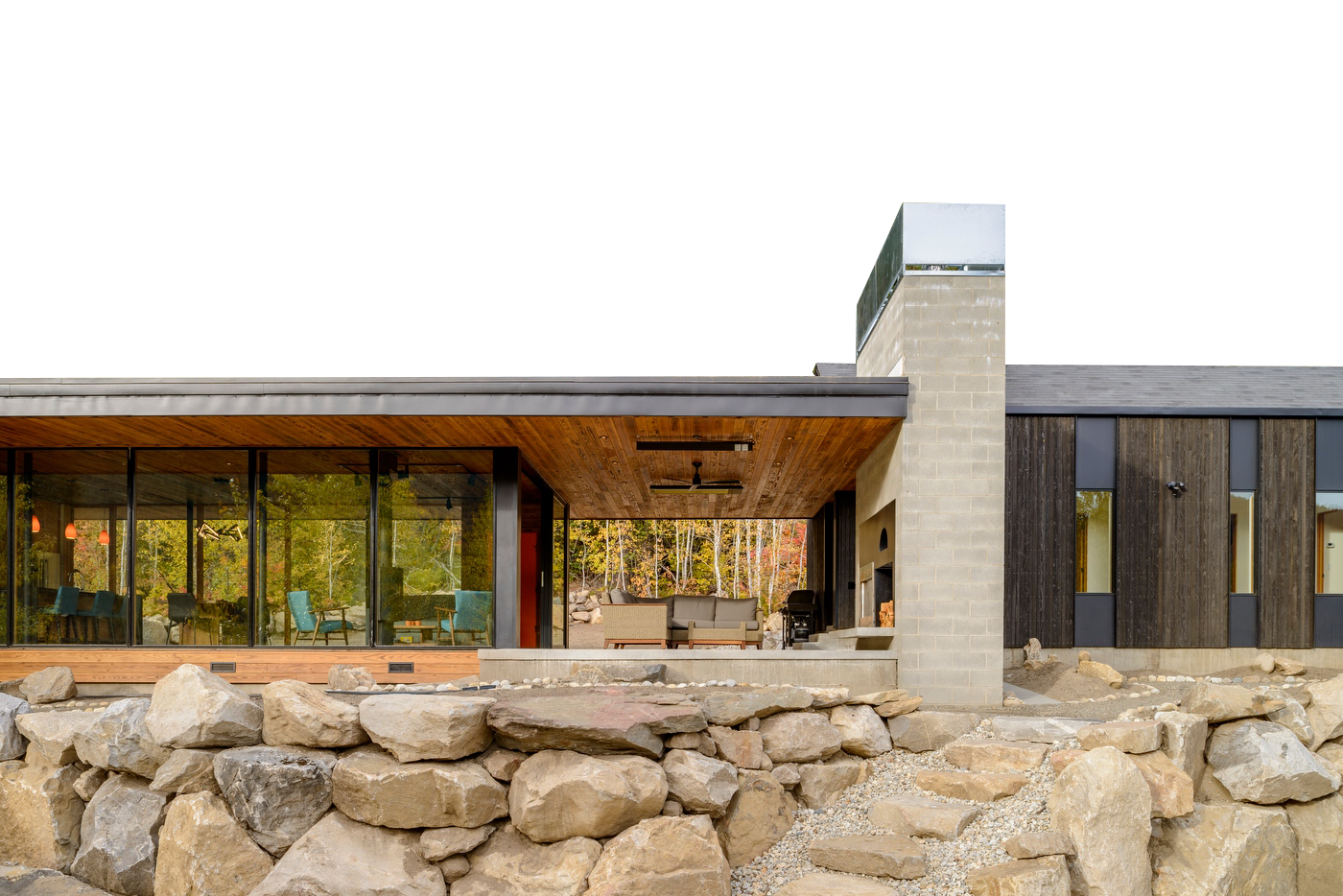 Modern house with a premium window and door company’s glass-walled living area, a covered outdoor patio, and a stone fireplace, built atop a natural rock foundation.