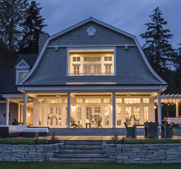 A two-story shingled home at twilight with a lit ground-floor porch featuring glass doors from a premium window and door company and patio furniture, overlooking a stone-walled lawn.