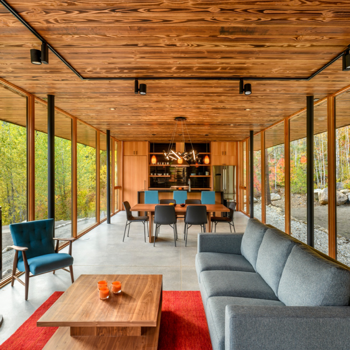 A modern living and dining room with premium window and door company installations, floor-to-ceiling windows, a wood-paneled ceiling, a grey sofa, a teal armchair, and a dining table.