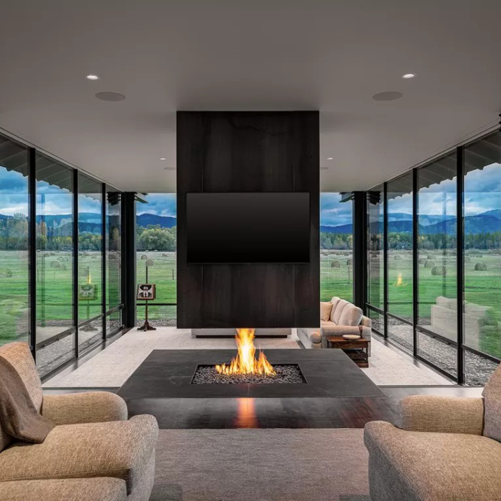 A living room featuring a double-sided fireplace and a television mounted on a central dark pillar, flanked by floor-to-ceiling windows installed by premium window and door services that look out over a grassy field.