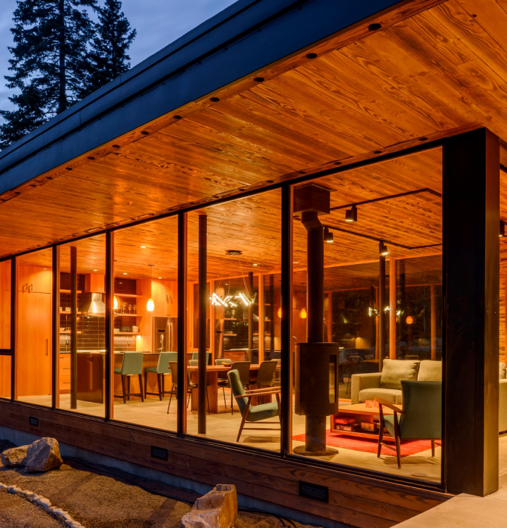 Modern home with floor-to-ceiling premium window and door services revealing an illuminated interior featuring an open-concept kitchen, dining area, and living space with a wood-burning stove.