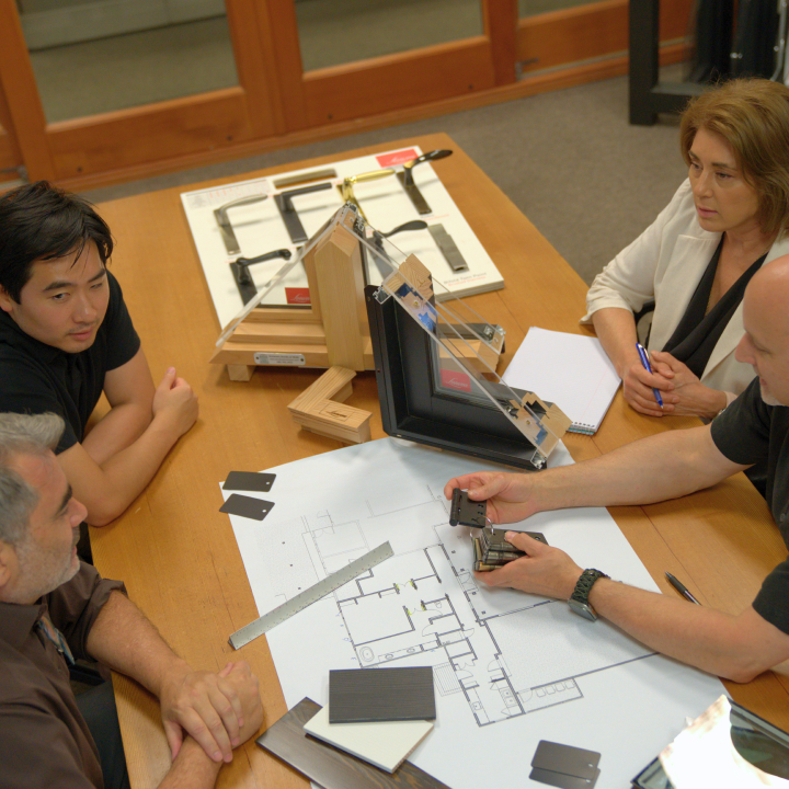 Four people sit around a wooden table reviewing architectural floor plans and window hardware samples for premium window and door services.