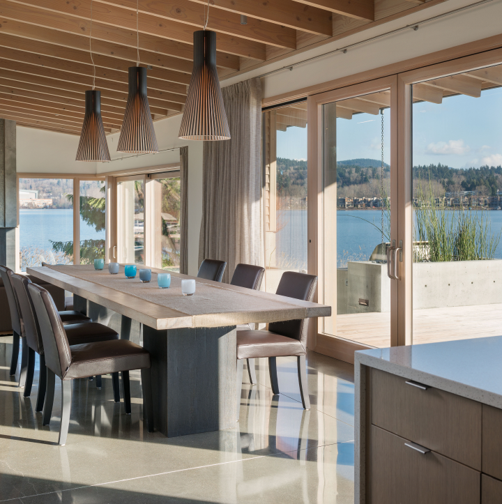 Dining room with a long wooden table, dark chairs, and three cone-shaped pendant lights, overlooking a lake through premium window and door services that frame the floor-to-ceiling views.