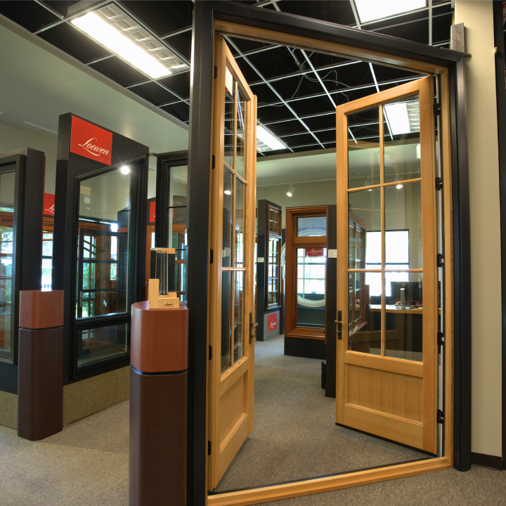 A showroom display featuring a pair of open wood-framed French doors and various other window and door samples labeled with Loewen branding, showcasing premium window and door services.