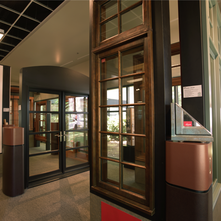 A showroom display of windows, doors and more featuring dark-framed glass doors and a multi-pane wooden window, with a metal corner sample resting on a pedestal in the foreground.