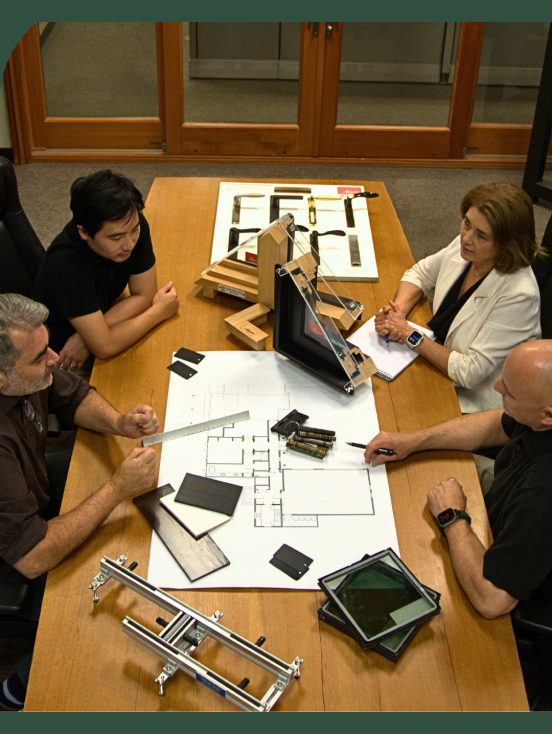 Four people sit around a wooden conference table in a windows, doors and more showroom examining architectural blueprints and construction material samples.