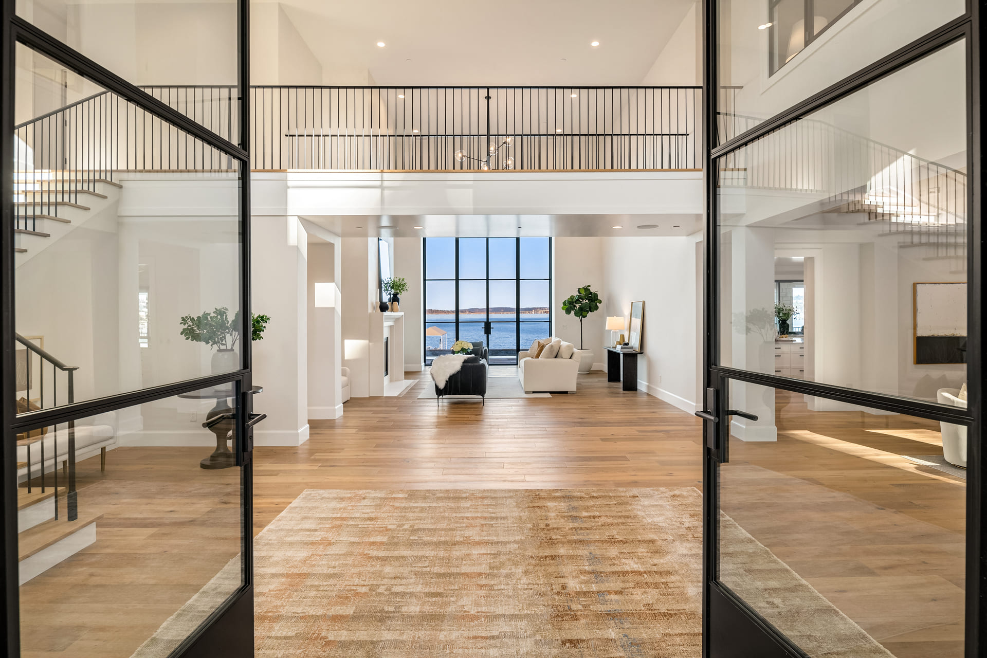 Open-concept living area with wood floors, a lofted balcony, and floor-to-ceiling glass from a premium window and door company, showcasing two black-framed doors that open to a view of the water.