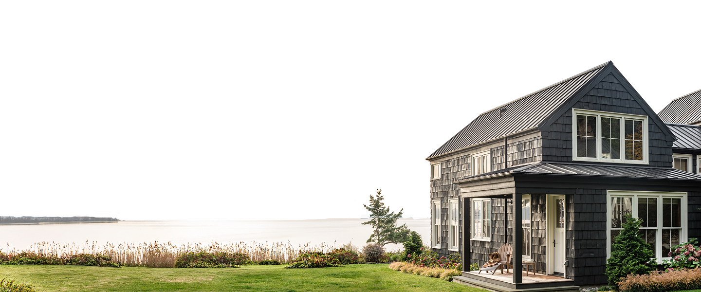 A dark-shingled house with a metal roof and Windows Doors and More brand features stands on a grassy lawn overlooking a calm body of water.