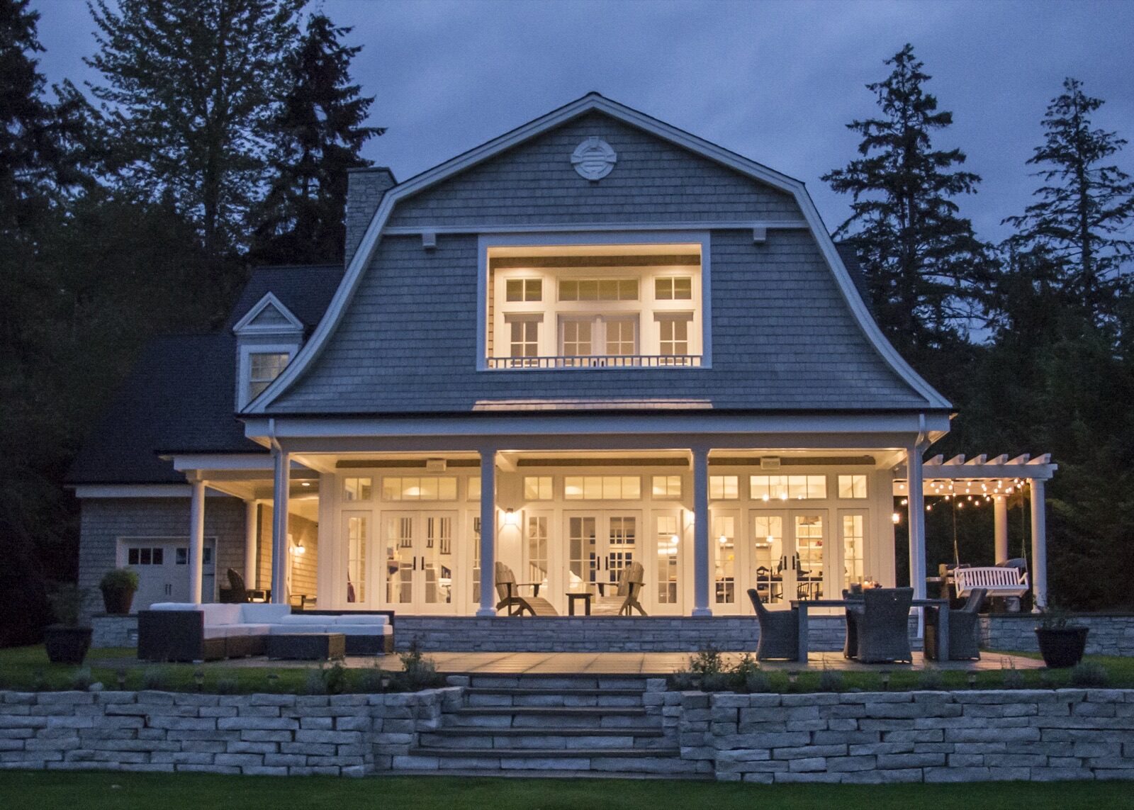 A two-story Andersen house with an illuminated ground-floor patio, outdoor furniture, and a stone terrace at twilight.