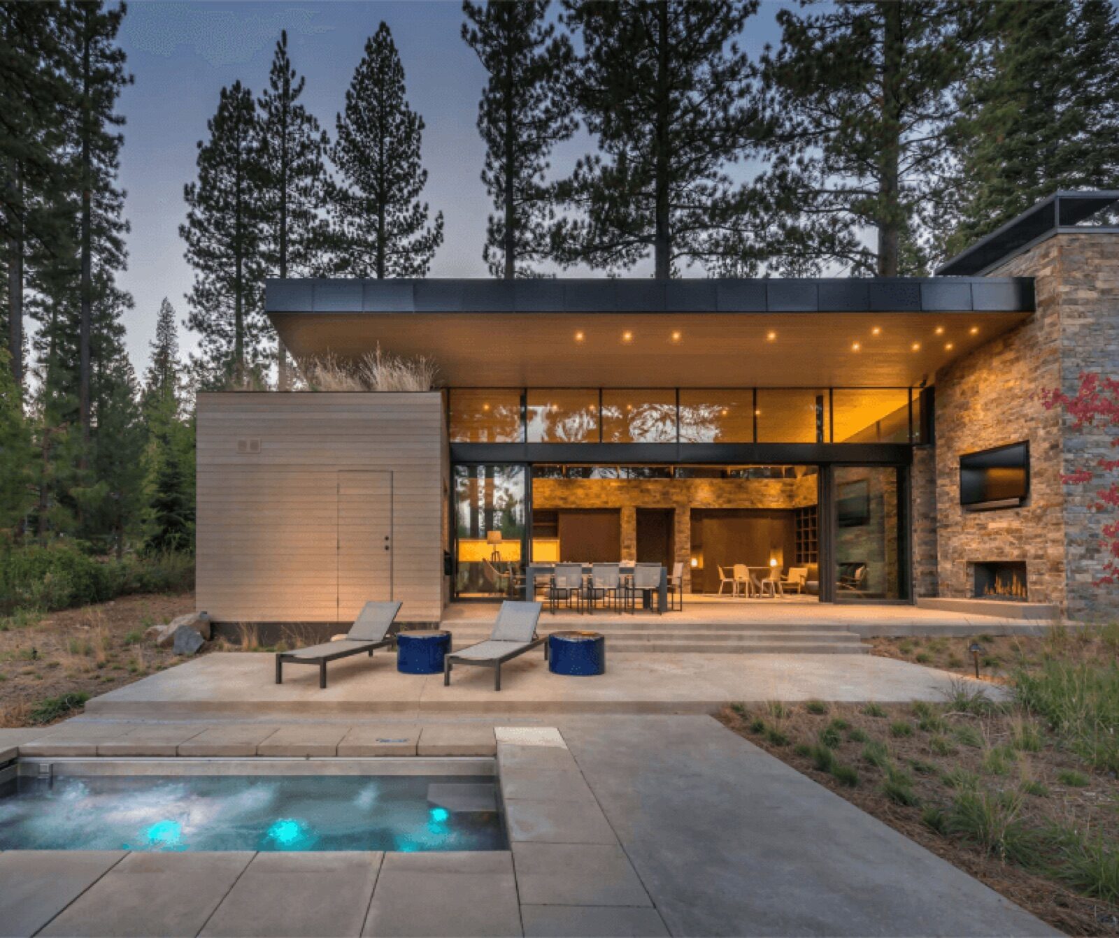 An Arcadia custom modern home with a flat roof, large glass walls, and an outdoor patio featuring a hot tub and lounge chairs, nestled in a forest at dusk.