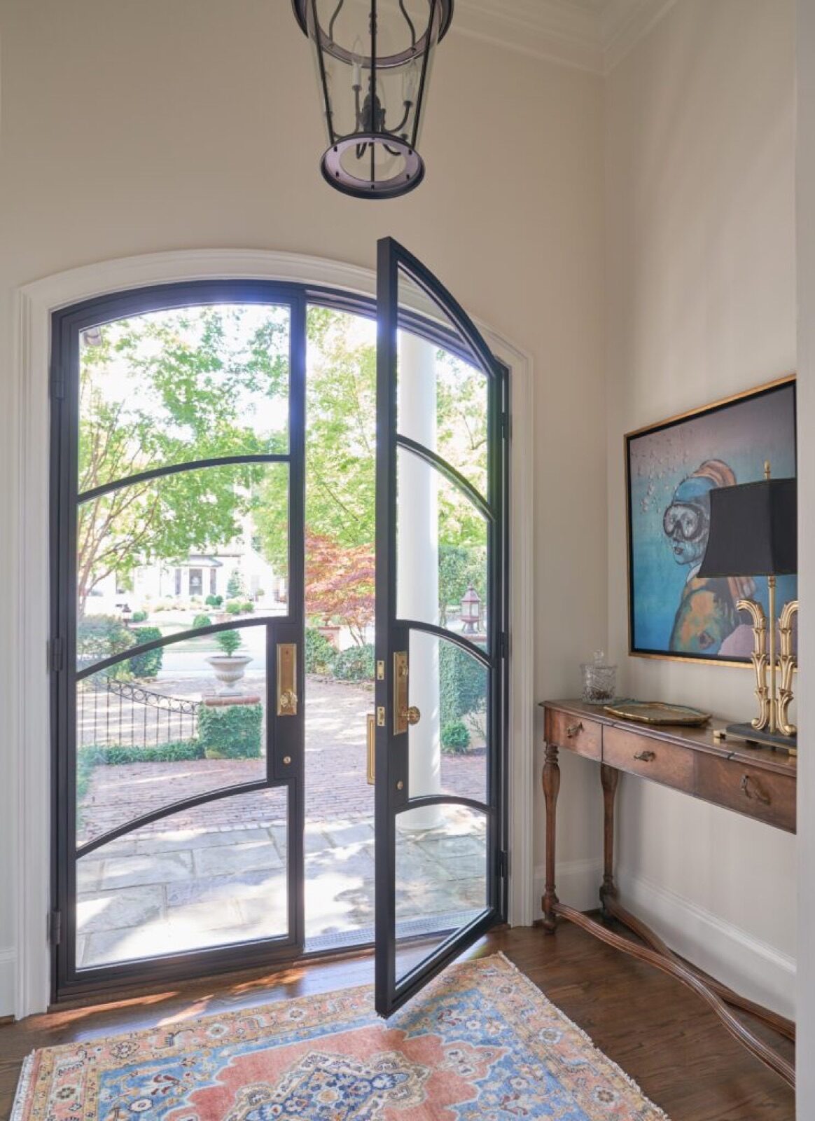 A glass-paned arched Clark Hall iron double door opening to an outdoor courtyard, with a console table and framed artwork in the entryway.