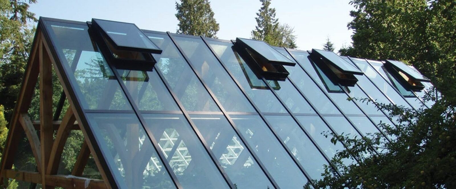 A sloped crystalite greenhouse roof with several open ventilation panels, framed by trees.