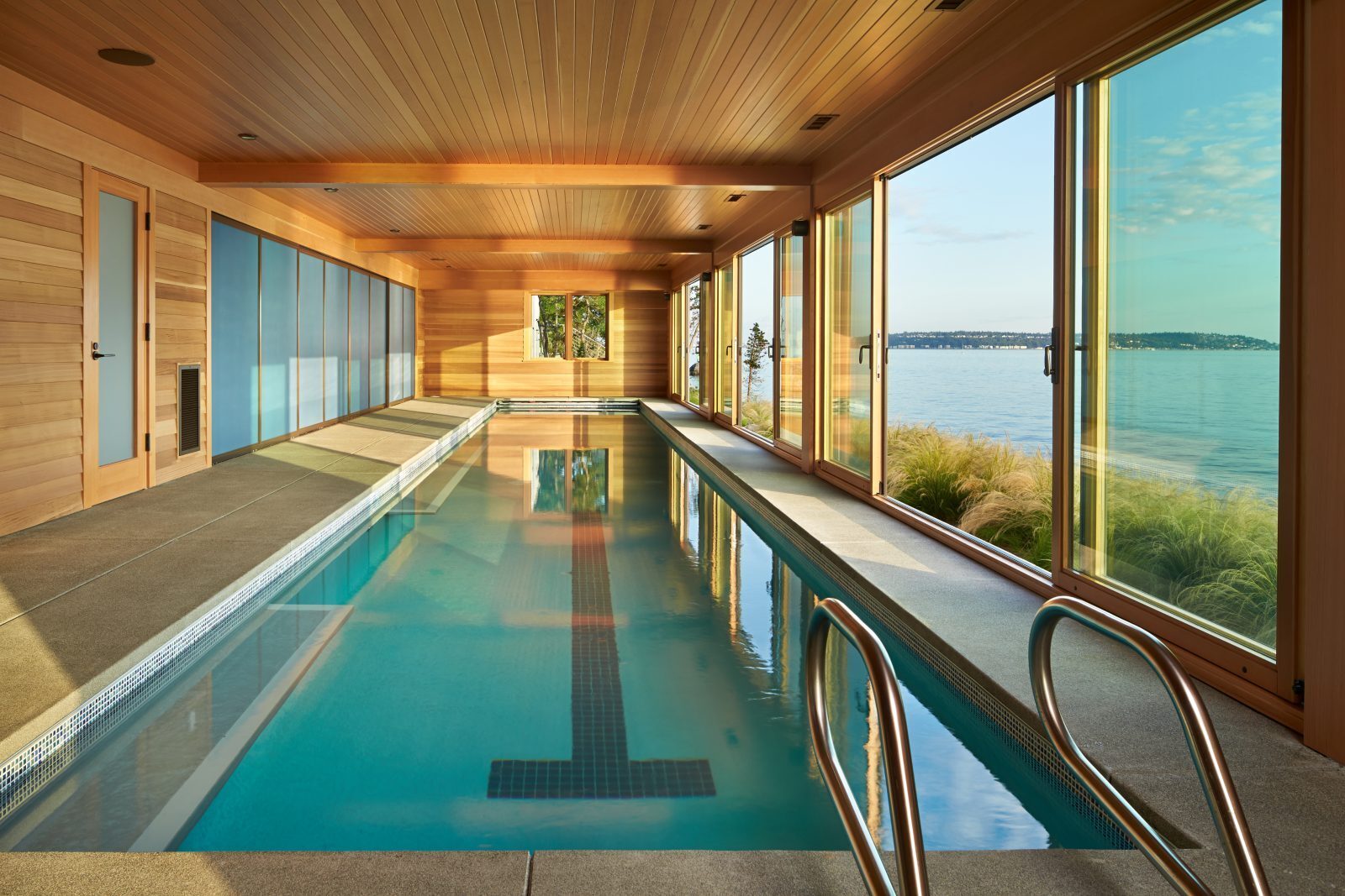 Indoor lap pool in a wood-paneled room with windows doors and more affiliates sliding glass walls overlooking the water.