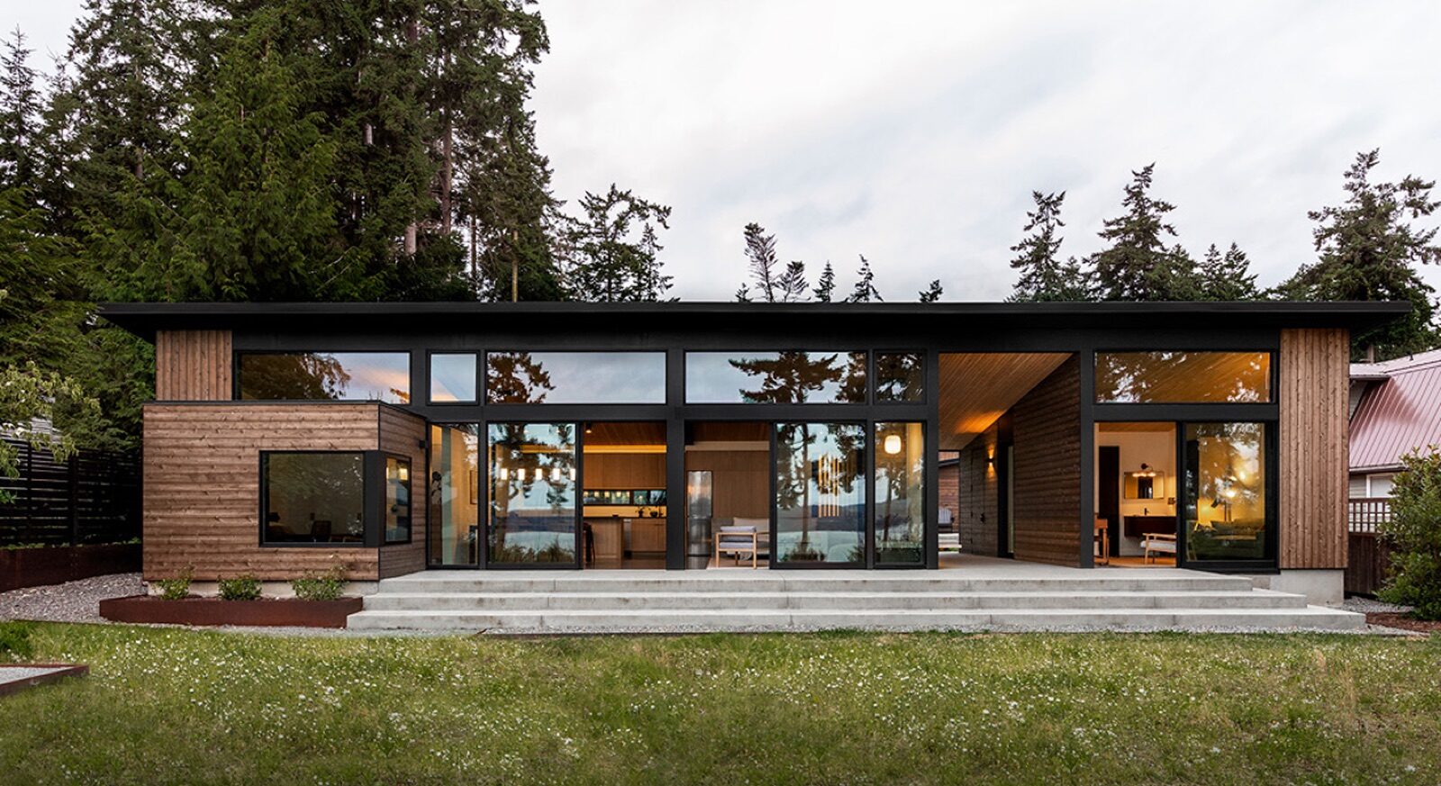 A modern, single-story home with horizontal wood siding and expansive Fleetwood floor-to-ceiling windows, featuring a concrete patio overlooking a grassy lawn.