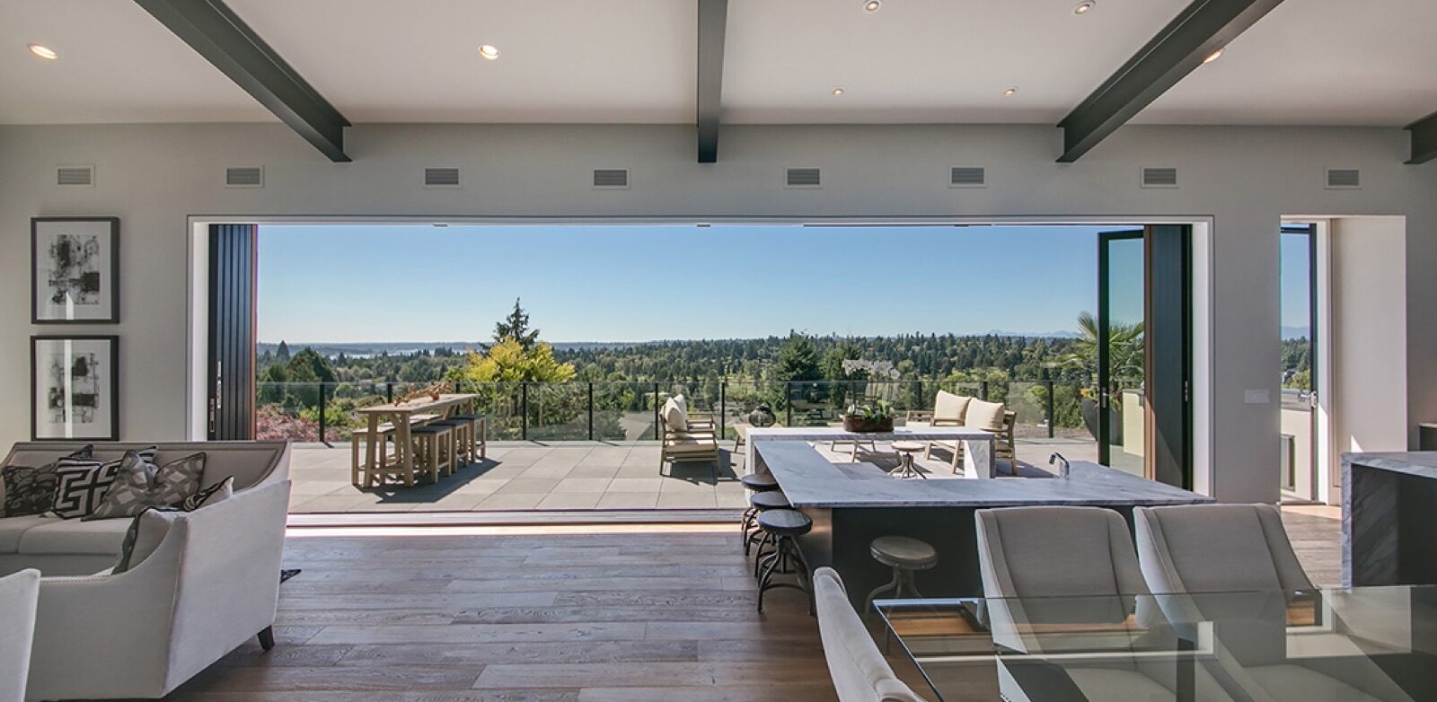 An open-concept living area with large LaCantina doors opening onto a patio overlooking a forest and distant water.