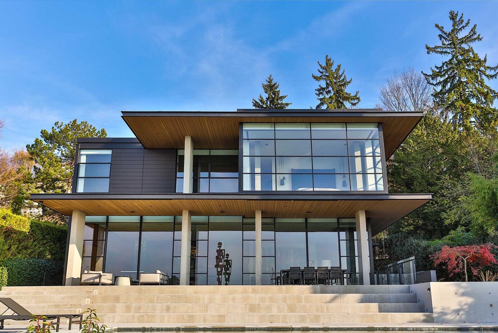 A modern mhb two-story home with large glass walls, flat overhanging roofs, and a concrete patio with outdoor seating.