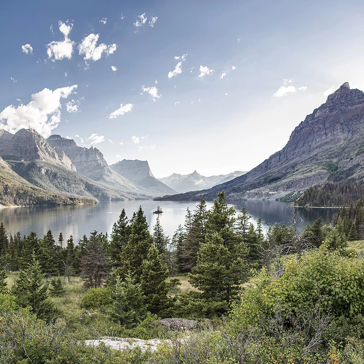 A wide mountain lake surrounded by pine forests and high, rocky peaks under a blue sky, reflecting the vistas seen through windows doors and more affiliates.