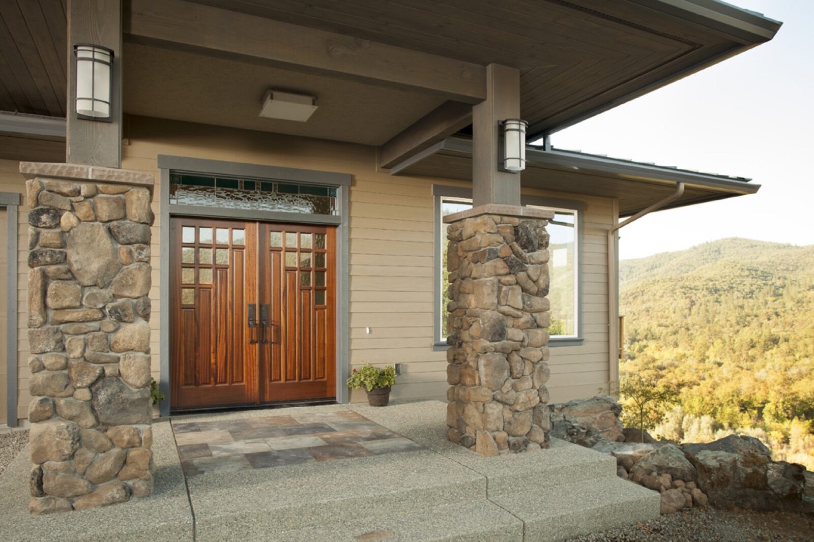 The front entrance of a house features Rogue Valley doors, stone pillars, and a view of forested hills.