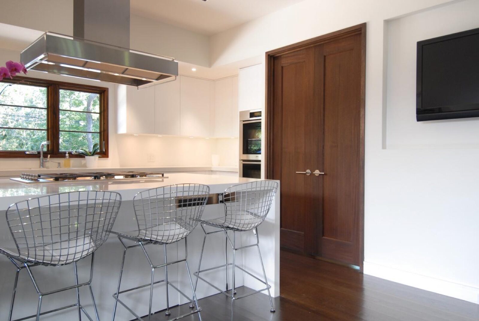 A modern kitchen with a white island and three wire-frame stools, featuring Trustile double doors and a wall-mounted television.