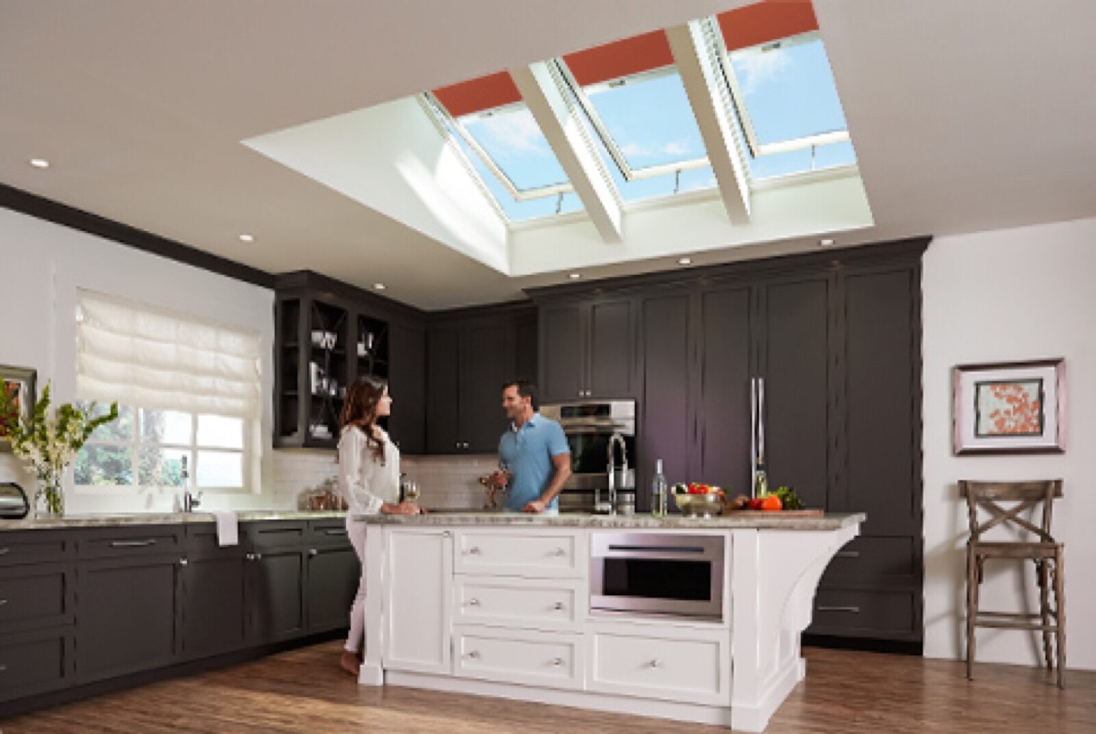 A couple stands in a kitchen featuring dark cabinets, a white center island, and a trio of Velux skylights in the ceiling.
