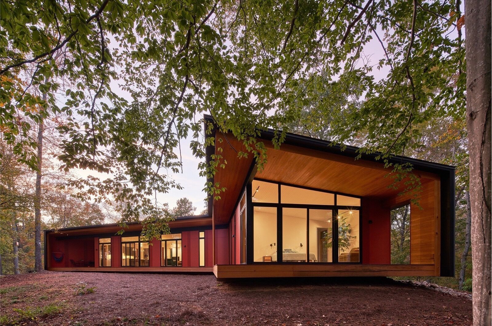 A modern red Weathershield house with a large wood-accented porch and floor-to-ceiling windows surrounded by trees.
