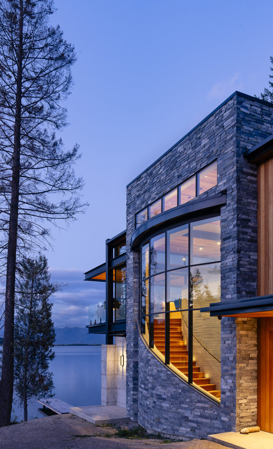 A modern stone-clad house by a lake at twilight, featuring an Awake Window and Door Co. curved glass wall that reveals an illuminated wooden staircase inside.