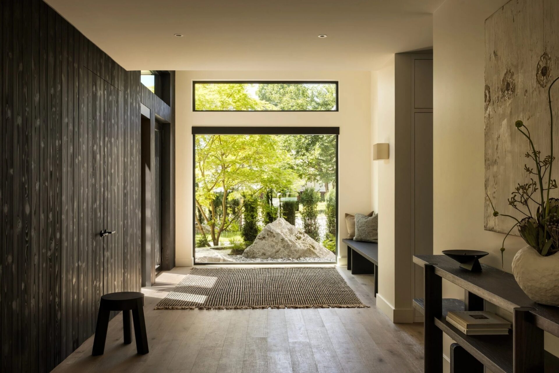 Modern entryway with light wood flooring, a striped rug, a small stool, and a large Euroline window looking out onto a garden with a large stone.