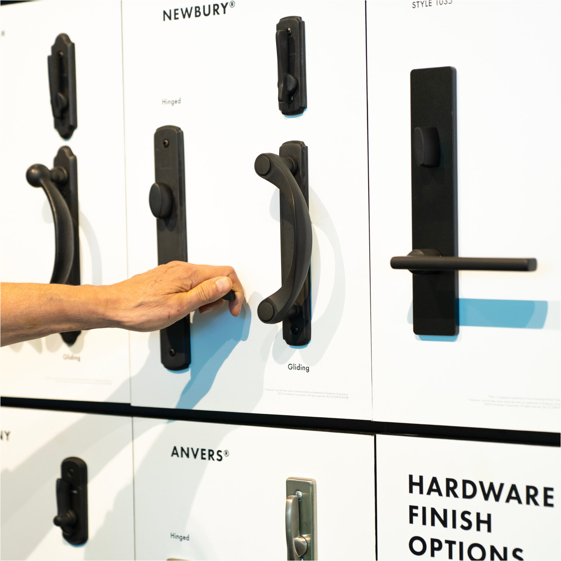 A hand tests the handle of a black gliding door lock on a windows doors and more showroom display board of various door hardware options.