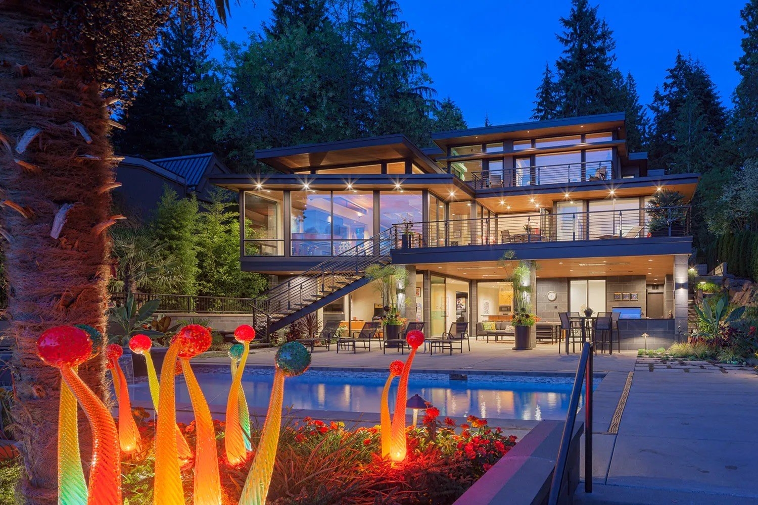 Modern Puget Sound home with expansive glass windows and a swimming pool, featuring illuminated glass garden sculptures in the foreground at dusk.