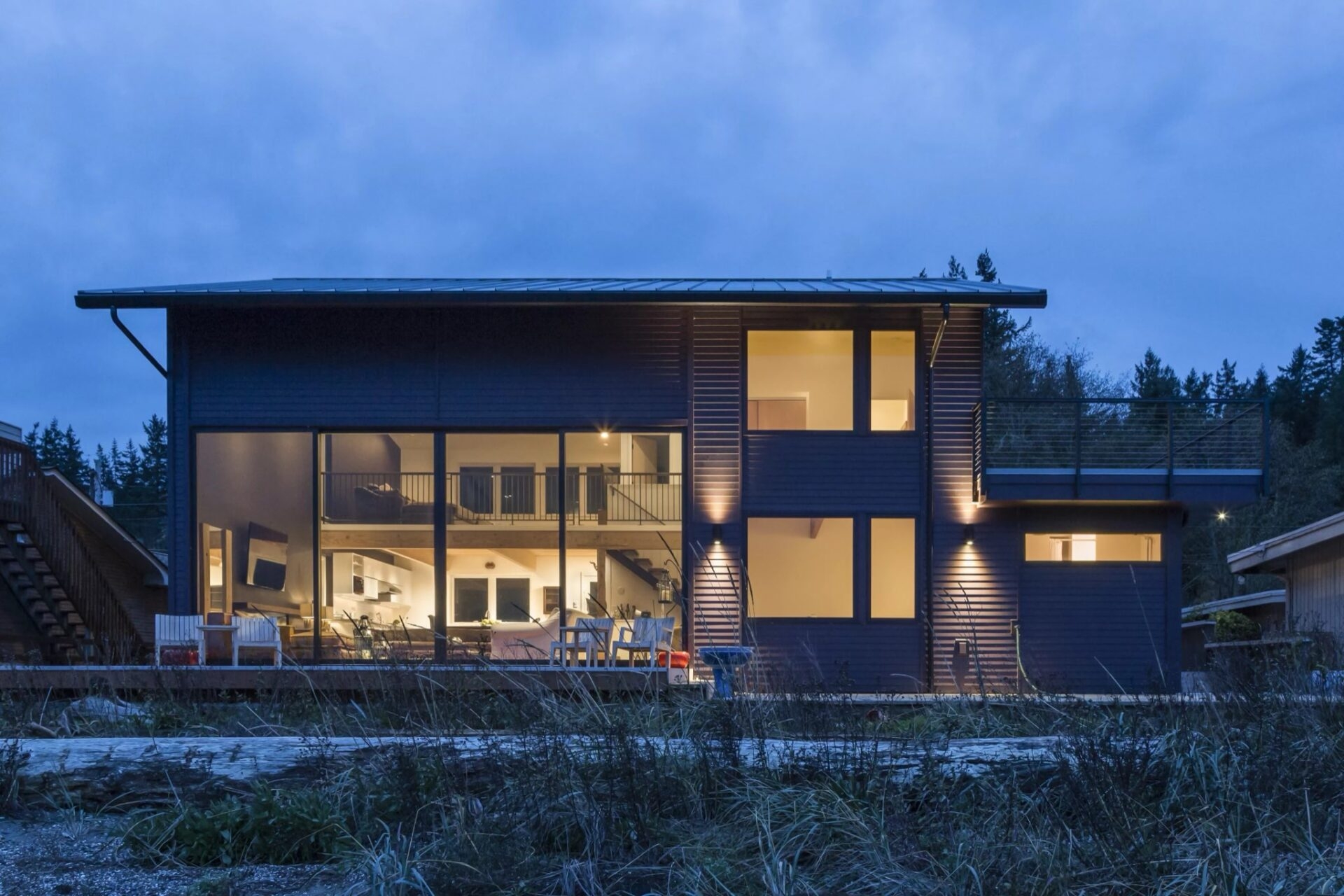 A two-story modern island beach house at twilight with large floor-to-ceiling windows revealing a lit interior and a balcony.