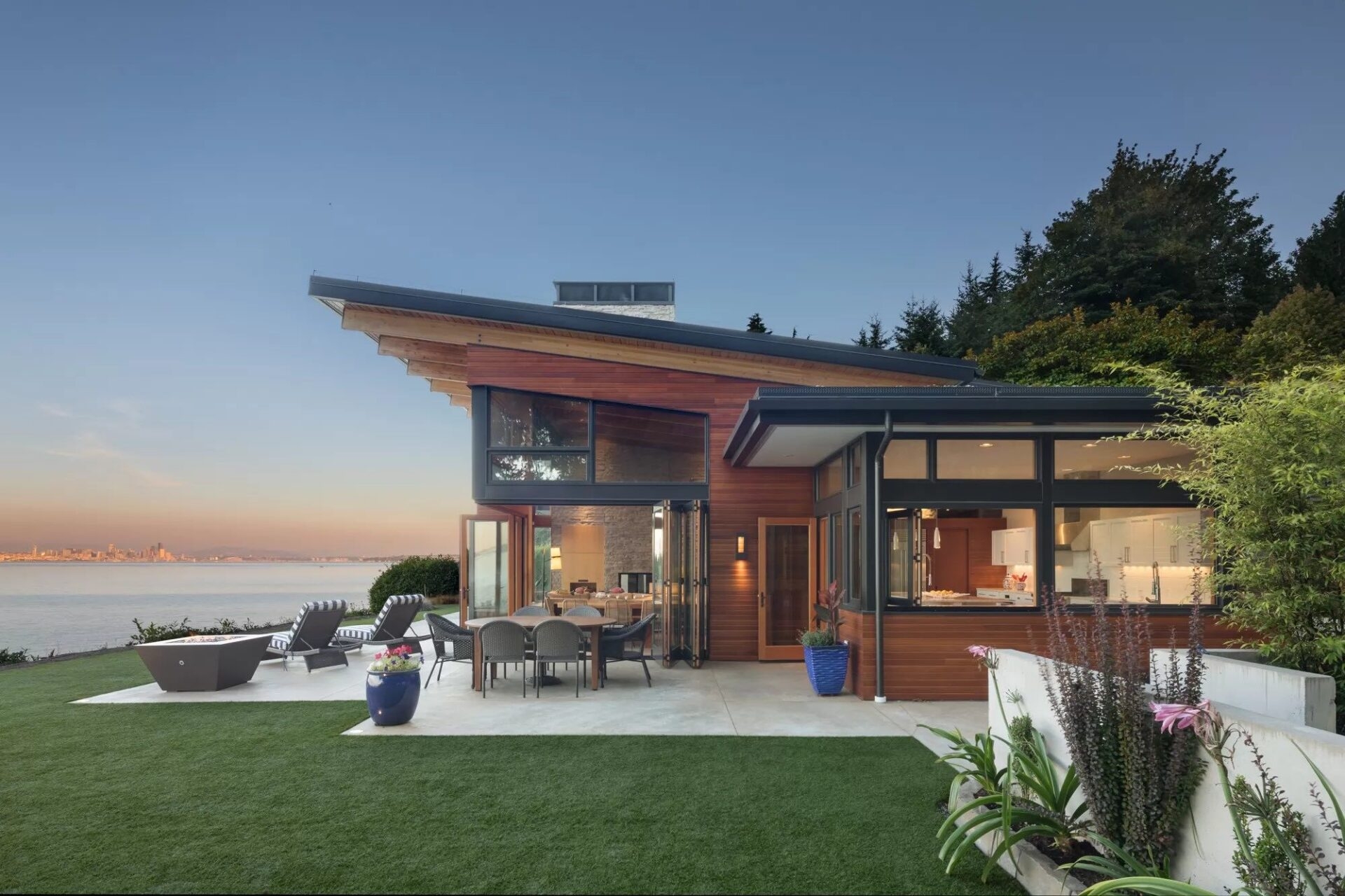A modern Bainbridge Beach House with a sloped roof, an open-concept living area, and an outdoor patio featuring a dining set and fire pit overlooking the ocean.