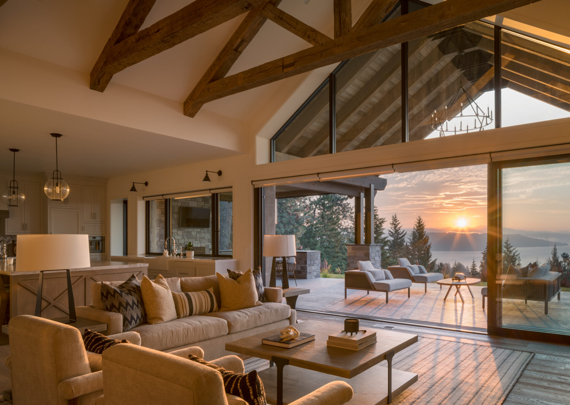 Living room with a beige sofa and armchairs opening onto a Loewen terrace with lounge chairs at sunset overlooking a lake.