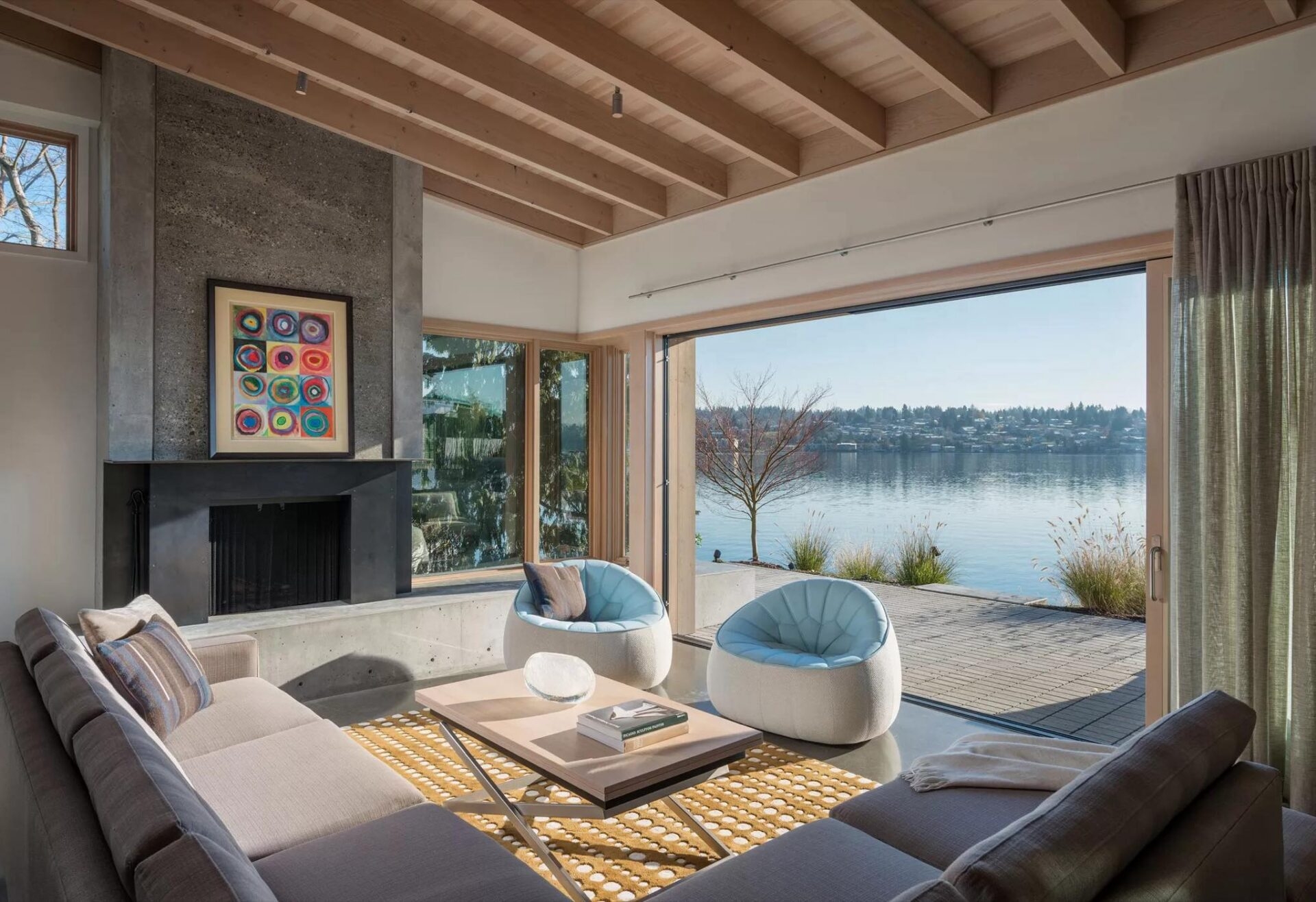 A Mercer Island contemporary living room with a concrete fireplace, colorful art, rounded light-blue chairs, and a sectional sofa facing a sliding glass door that opens to a waterfront view.