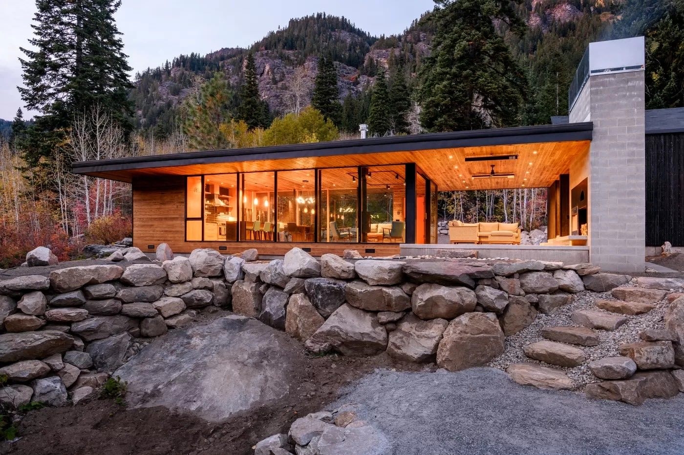 Modern River House with floor-to-ceiling windows and a covered patio, nestled in a mountainous landscape behind a large stone retaining wall.