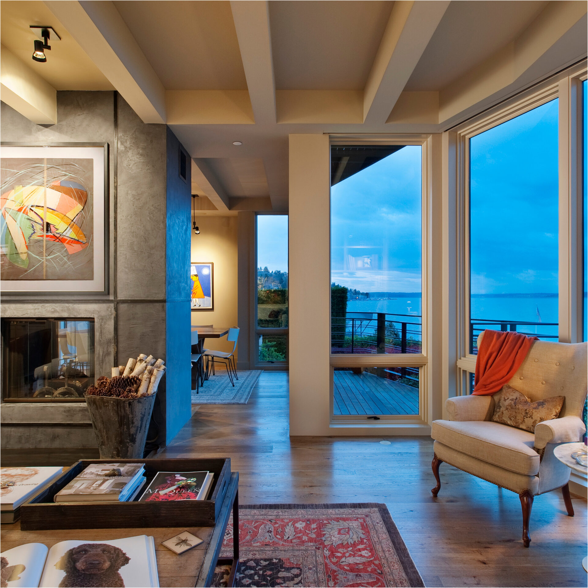 A cozy living room with a fireplace, a beige armchair with an orange throw blanket, and floor-to-ceiling windows from a premium window and door company overlooking a dark blue lake.