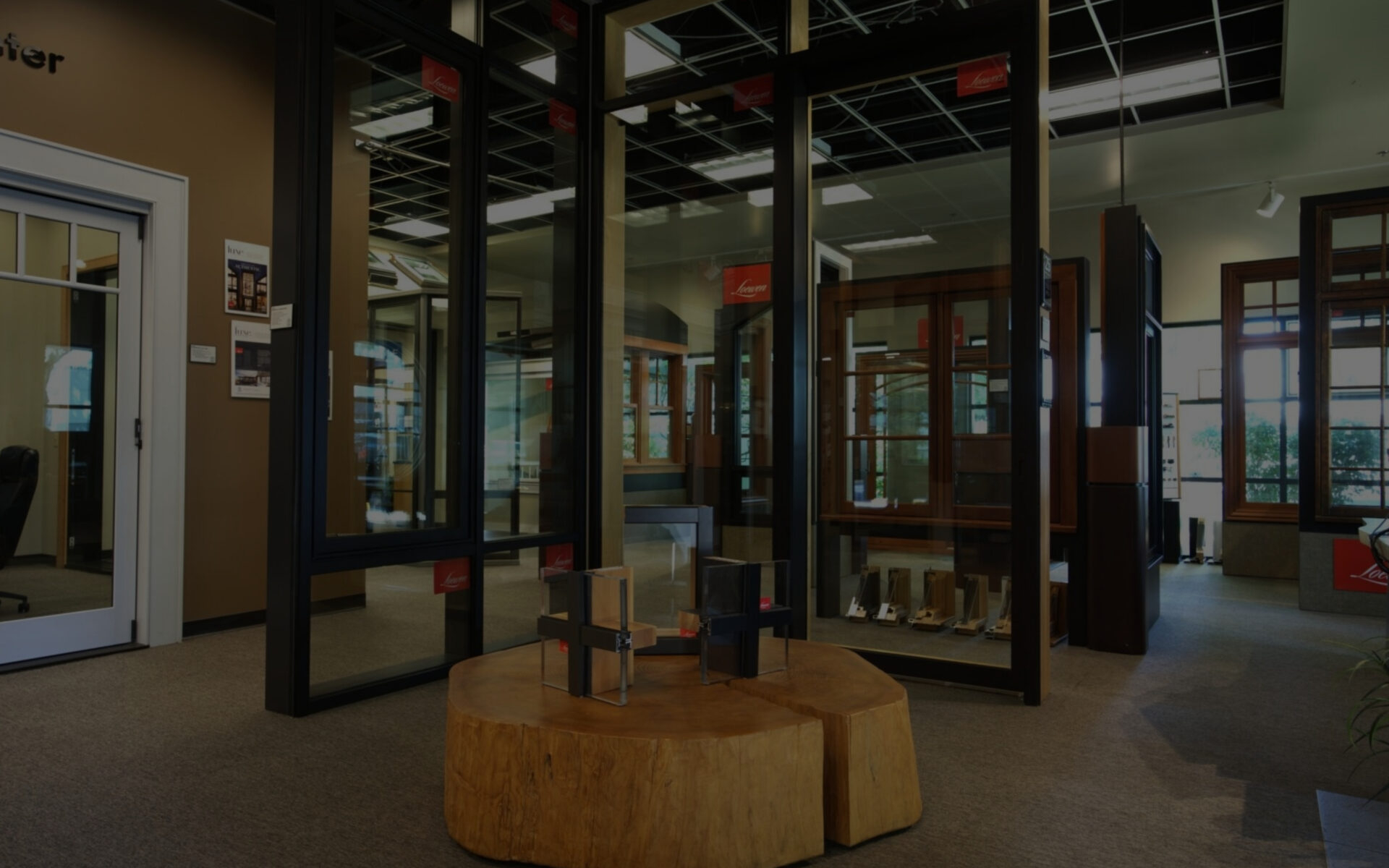 A windows doors and more showroom displaying various window and door frames, with a wooden stump table in the foreground holding small product cross-sections.