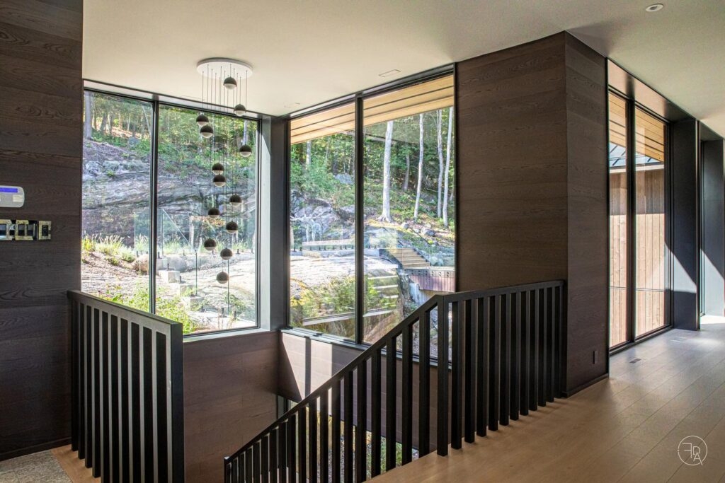 A modern staircase with black railings beside floor-to-ceiling western architectural openings looking out onto a wooded, rocky hillside.
