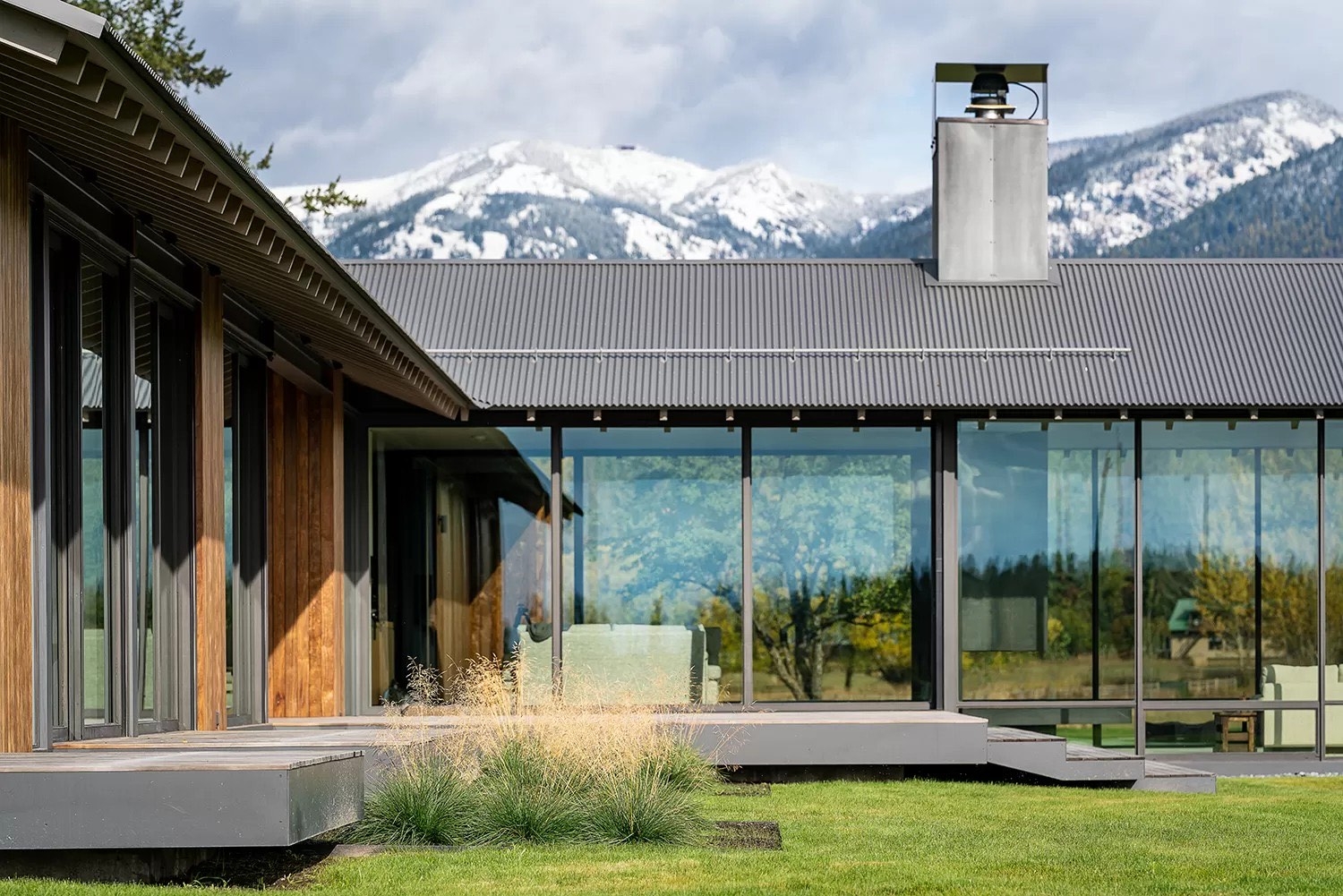 Whitefish modern house with a metal roof and large glass windows overlooking a grassy yard and snow-capped mountains.