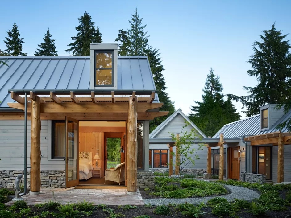 Modern cabin with a metal roof and log posts, serving as a coastal getaway. An open doorway reveals a bedroom interior, and a stone path leads to a connected structure nestled among pine trees.