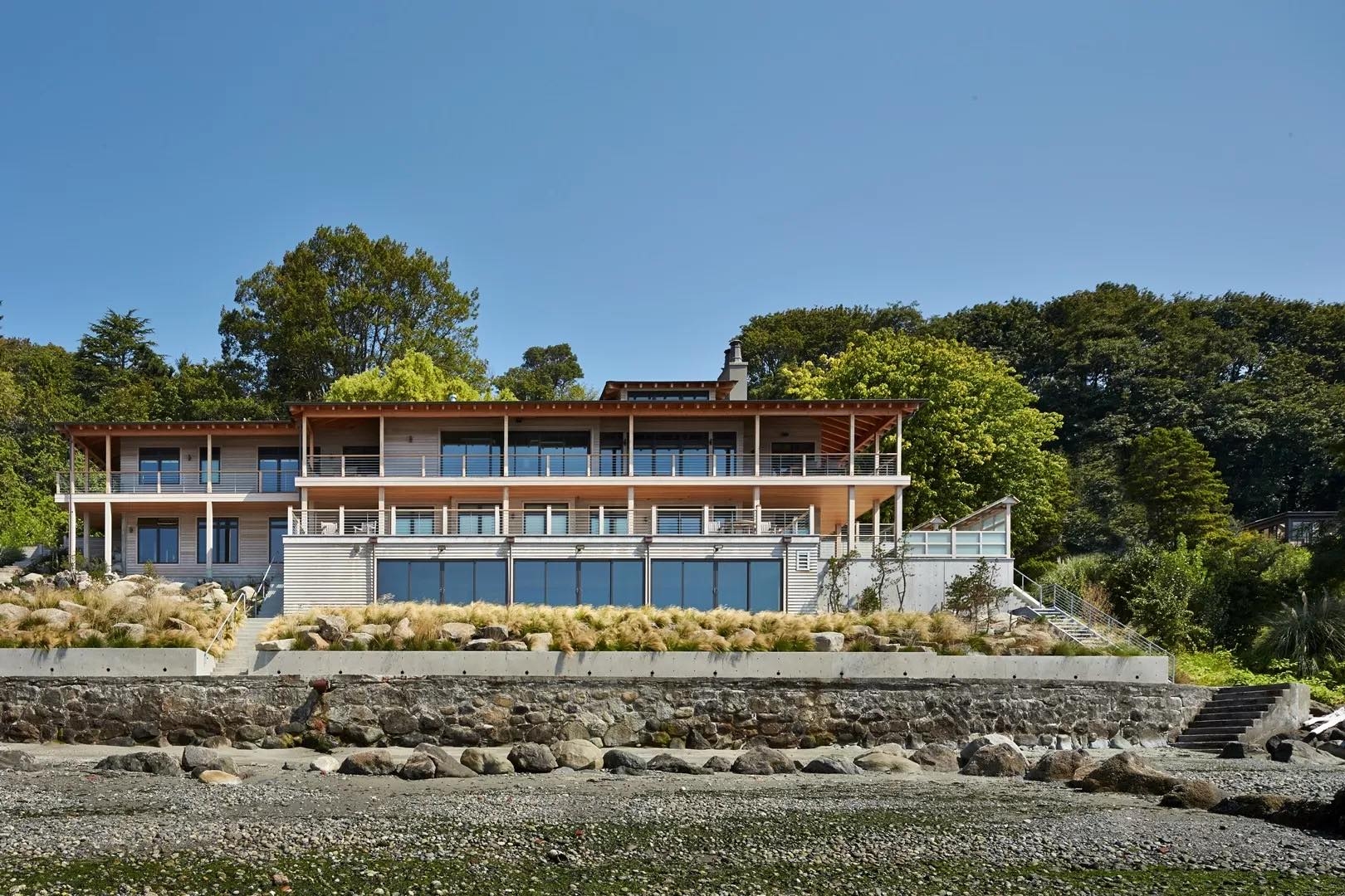 A modern three-story Harbor House with wrap-around balconies and large glass windows, situated on a stone terrace above a rocky beach.
